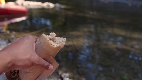 mano sosteniendo un sándwich medio comido sobre el arroyo mientras acampa