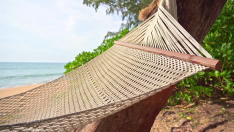 Nahaufnahme-Einer-Leeren-Gewebten-Hängematte,-Die-Im-Wind-Schaukelt,-Wellen,-Die-An-Einem-Sandstrand-Im-Hintergrund-Hereinrollen