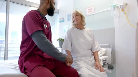 diverse male doctor holding hand of senior female patient on bed in hospital room, slow motion