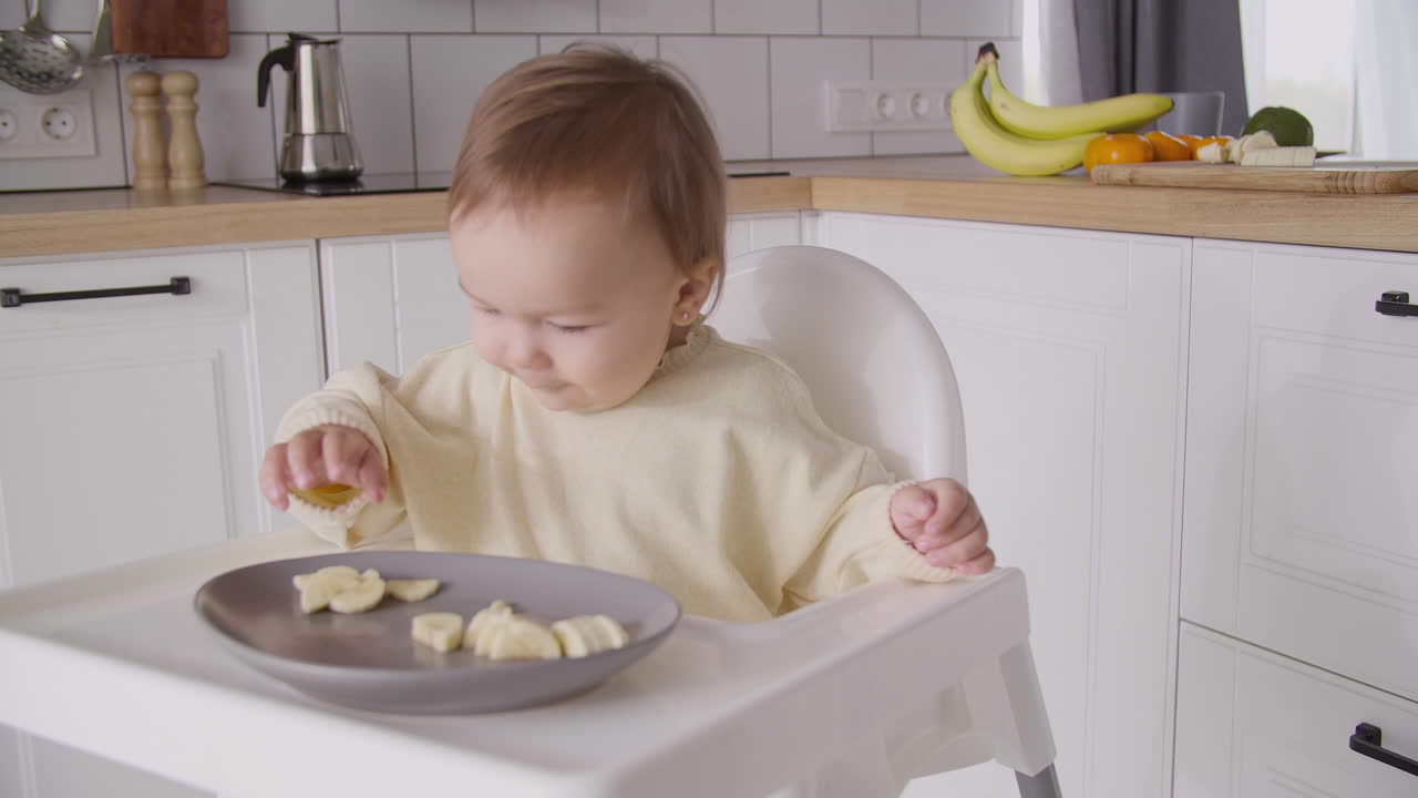linda niña comiendo rodajas de plátano sentada en su silla alta en la cocina 7