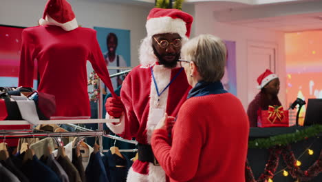 Empleado-Vestido-Con-Traje-De-Papá-Noel-Y-Gafas-Ayudando-A-Un-Cliente-Anciano-En-Una-Tienda-A-Encontrar-La-Blusa-Roja-Necesaria-Para-El-Atuendo-De-Fiesta-De-Navidad.-Trabajador-Ayuda-A-Una-Mujer-Mayor-En-Una-Tienda-De-Moda-Durante-Las-Vacaciones-De-Navidad