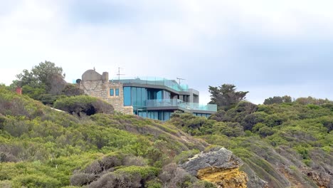 A modern house with glass facade sits atop lush green hills, under a cloudy sky, along Australia's Great Ocean Road