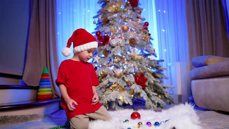 little kid in santa cap has the toys on the fluffy plaid. toddler shows the decorations to the camera. christmas tree at backdrop.