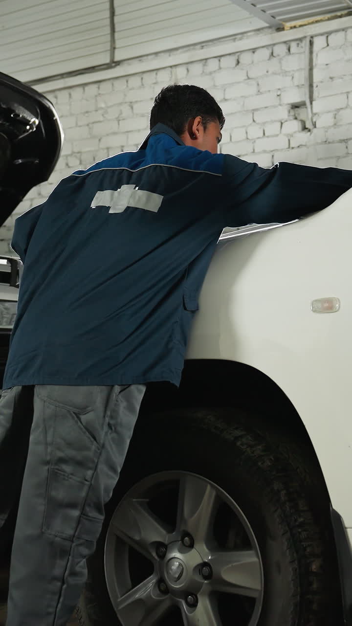 mecánico en uniforme azul revisa el neumático del coche golpeándolo con la pierna en el taller de automóviles, realizando la inspección y mantenimiento del vehículo en un centro de servicio mecánico