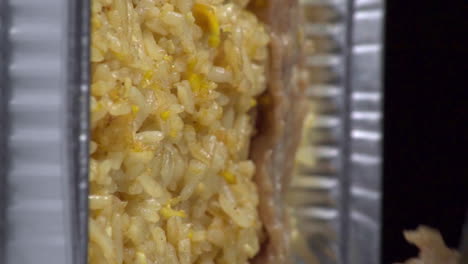 dutch angle vertical view of cooked slices of meat being added and layered above yellow rice surface with tongs in silver foil take out pan, static extreme close up detail