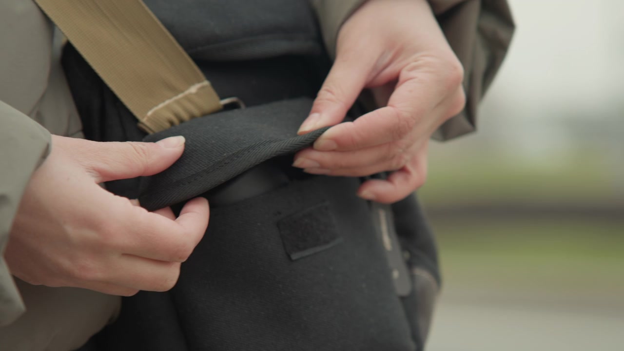 Hands opening black canvas pouch outdoors, closeup showing gentle finger movement on flap, khaki strap, textured fabric, muted city background and overcast light, practical retrieval of small camera