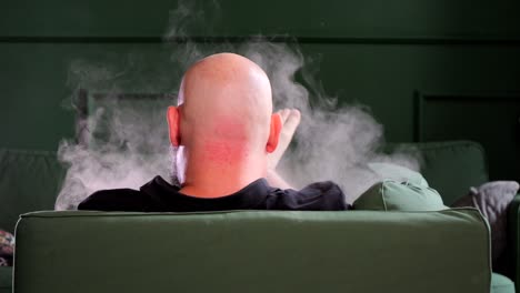 un hombre calvo relajándose en un sofá verde usando un cigarrillo electrónico