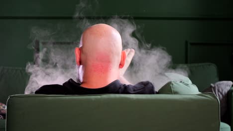 a bald man relaxing on a green couch using an e-cigarette