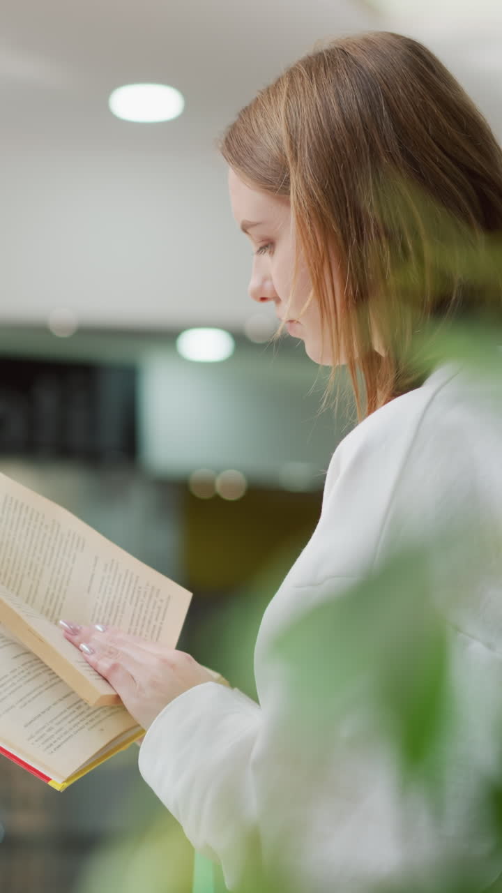 una mujer joven hojeando un libro mientras está sentada en un moderno centro comercial rodeada de luz suave, reflejos de vidrio y vegetación vibrante