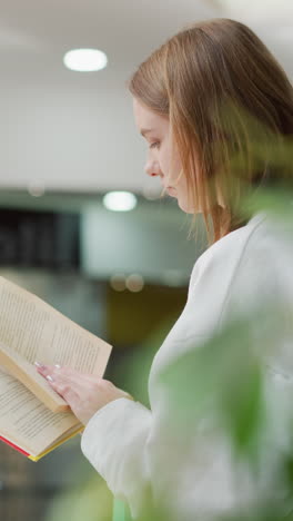 una mujer joven hojeando un libro mientras está sentada en un moderno centro comercial rodeada de luz suave, reflejos de vidrio y vegetación vibrante
