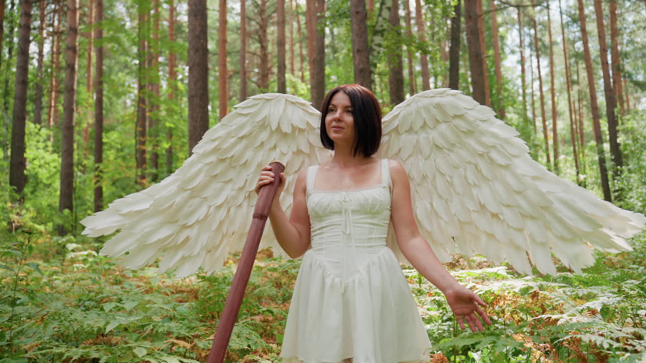 Young lady with white feather wings dressed in flowing white gown walking along forest path holding wooden staff gently touching green plant leaves as sunlight filters through trees