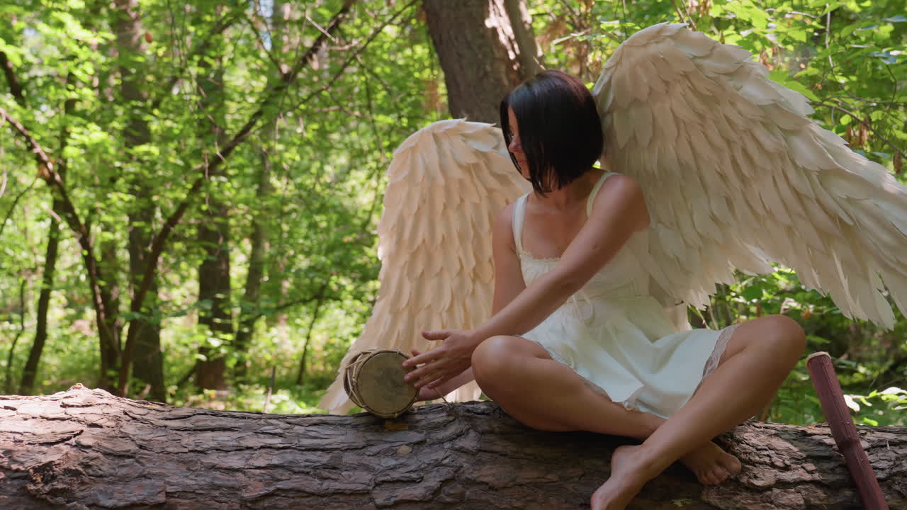 Fairy lady with large white wings sits on forest log holding traditional drum, sunlight shimmering through leaves, capturing calm mystical moment before she drops drum