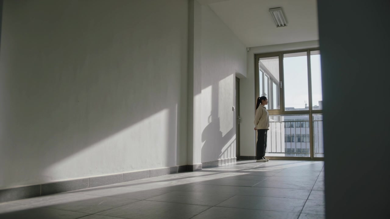 Mujer de pie con chaqueta clara y pantalones oscuros, mirando por la ventana, la luz del sol proyectando una sombra, espacio para texto.