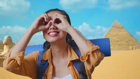 close up of asian female hiker with mountaineering backpack smiling and making binoculars gesture then looking around while traveling in pyramid of giza