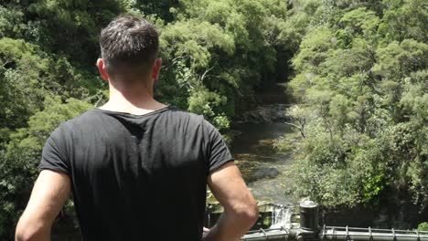 SLOWMO---Young-male-tourist-looking-down-at-Mokora-Falls-from-lookout,-Auckland,-New-Zealand