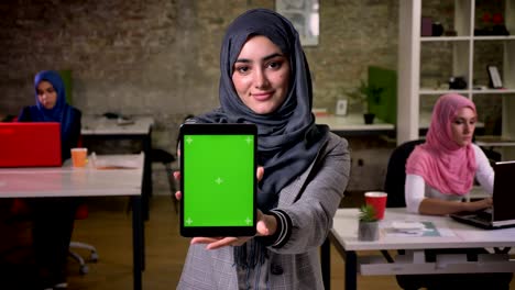 cute arab female standing and showing green screen on her tablet while standing chill with nice smile, colleagues on background, brick wall