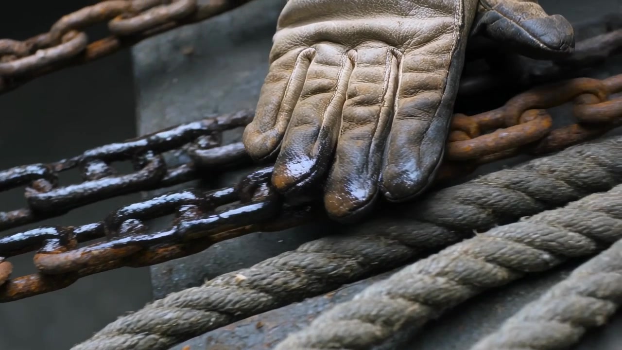 Mano guantata che tocca un anello oleoso, premendo e trascinando le punte su catena e corda sul ponte, sporcando il guanto.