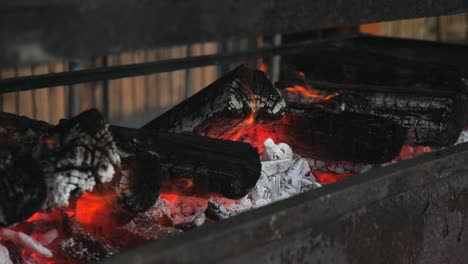 slow motion pan along metal barbecue with open orange hot fire flame flickering as ash and embers rise up from smoke with red and white charcoal bed in rustic outdoor setting