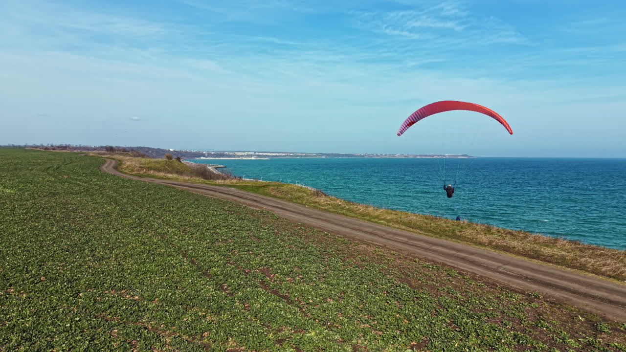 Pessoa praticando parapente sobre a costa em um dia claro perto do oceano, com campos ao fundo e uma linha costeira visível à distância.
