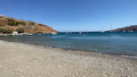 greece, astypalea island, livadi beach on a sunny day. people enjoying themselves by the beach, some people swimming and some sun bathing, sailboats anchored further on the deep blue sea.