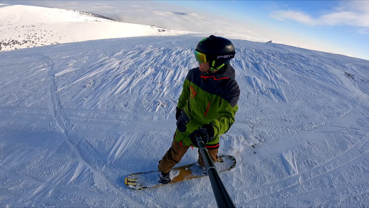 Professionally performed descend on the snowboard by the mountain slope. Sportsman in protective helmet and glasses takes selfie of the ride.