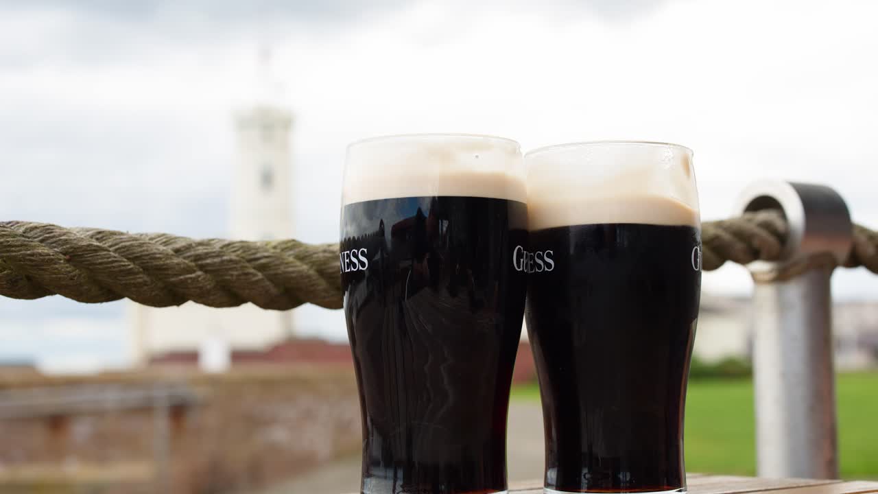 Two hands clink pints of dark beer outdoors by the water on a cloudy day