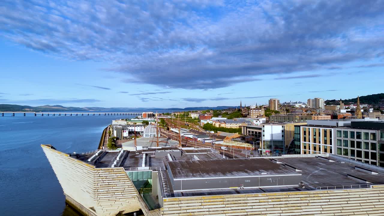Drone camera smoothly pans above a striking contemporary museum along Dundee’s waterfront, revealing cityscape, river, and dramatic morning sky in natural light