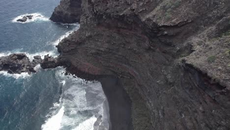 Ocean-Waves-Splashing-Onto-Shore-With-Black-Volcanic-Sand-And-Steep-Rugged-Cliff-At-Nogales-Beach-In-La-Palma,-Canary-Islands,-Spain