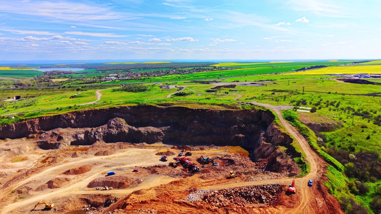 Grüne Hügel und sonniger Himmel. Blick von oben auf einen Steinbruch mit Maschinen, entferntes Ackerland und ein kleines Dorf.