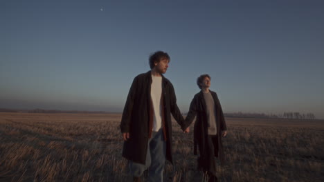 couple holding hands and walking through open field at sunset