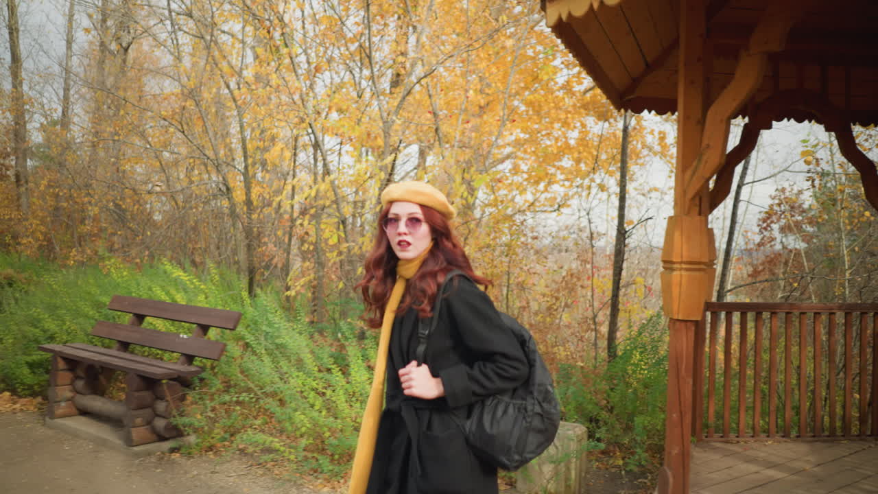 검은 코트, 노란 스카프, 베레트를 입은 관광객이 조용한 가을 공원에서 목조 게지보에서 나와, 집중된 표정으로 가방 줄을 들고, 배경에 목조 벤치를 들고 있습니다.