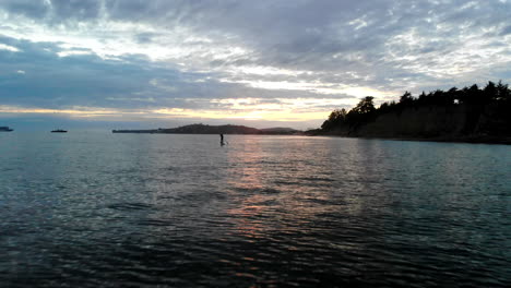 Drone-Aéreo-Disparado-En-Montecito-Al-Atardecer-Volando-Bajo-Sobre-El-Agua-Y-Reflejos-Del-Océano-Pacífico-Con-Un-Surfista-De-Remo-A-Lo-Largo-De-Los-Acantilados-De-La-Playa-De-California-En-Santa-Barbara,-California