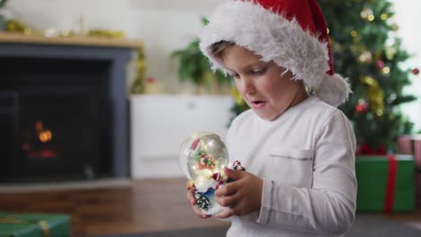 Überraschter-Kaukasischer-Junge-Mit-Weihnachtsmütze,-Der-Mit-Der-Schneekugel-Spielt