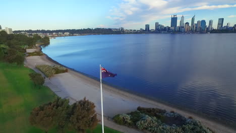 Inclinación-Aérea-Hacia-Abajo-Sobrevuelo-Del-Asta-De-La-Bandera-Australiana-En-El-Parque,-La-Ciudad-Y-El-Río-En-Segundo-Plano.