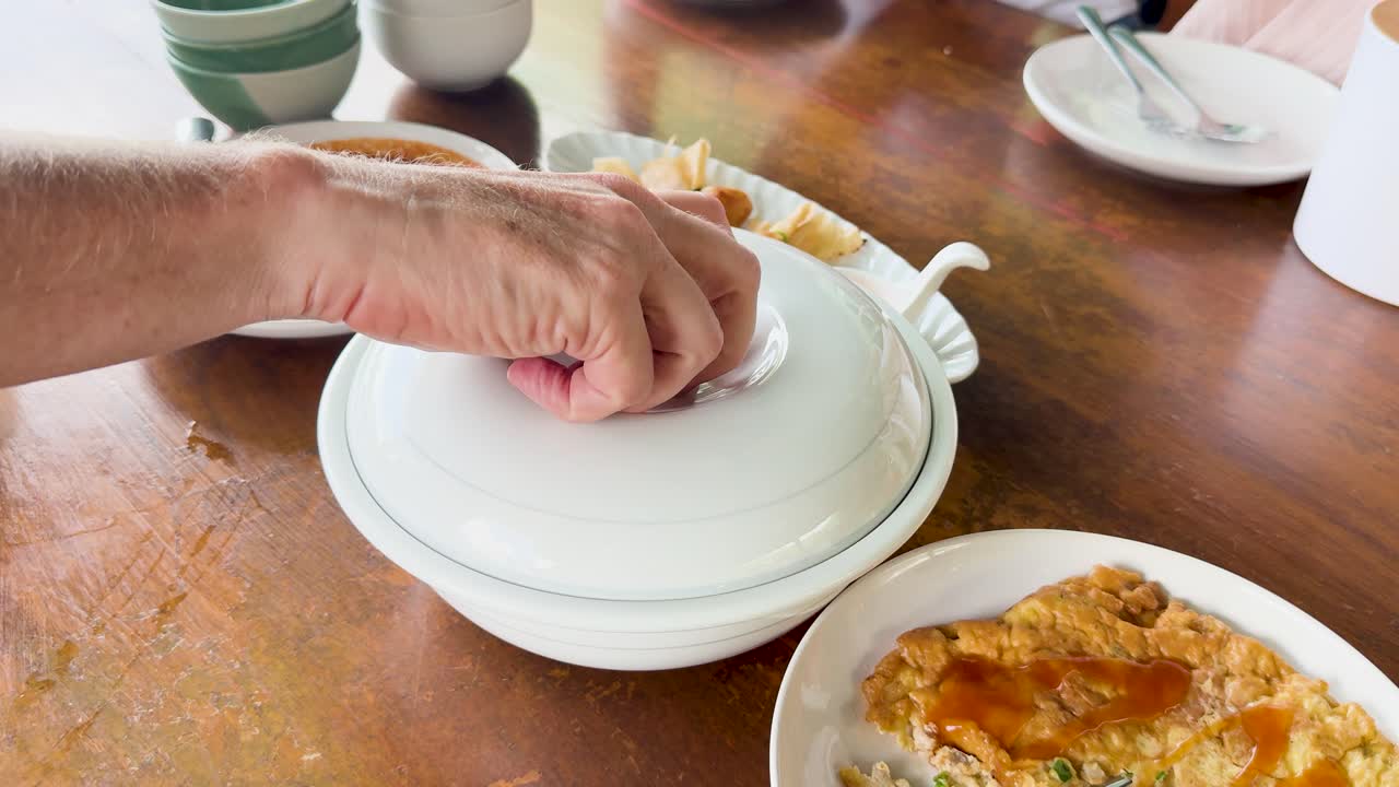 A hand uncovers steaming white rice at a clifftop restaurant in Phuket, Thailand. Bright natural lighting enhances the inviting dining scene