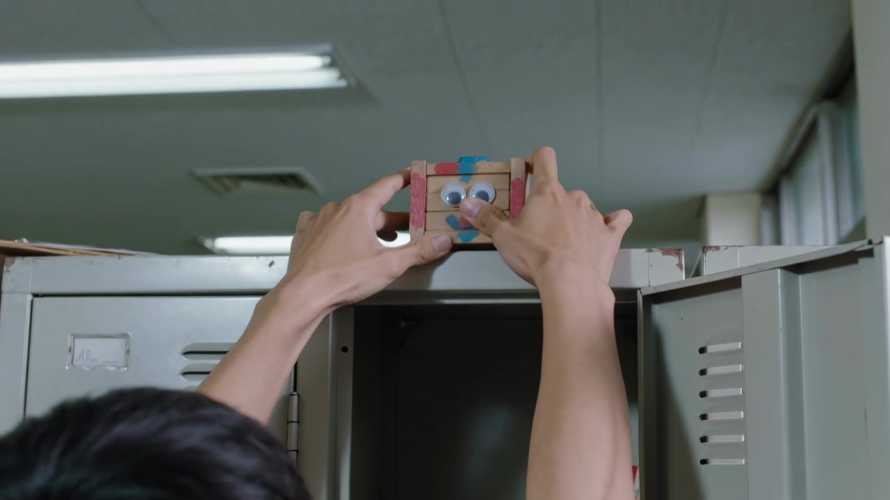 Opening locker, reaching teen removing wooden toy, placing googly-eyed box on locker top at school