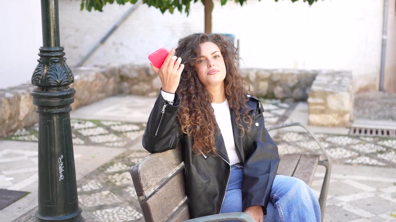 Una giovane donna elegante è seduta con grazia su una panchina del parco, tenendo uno smartphone alla luce del giorno.