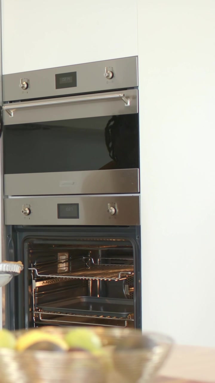 Video verticale: Uomo in maglietta scura e guanti da forno che apre il forno inferiore, toglie una teglia e la appoggia sul bancone, con spazio per il testo.