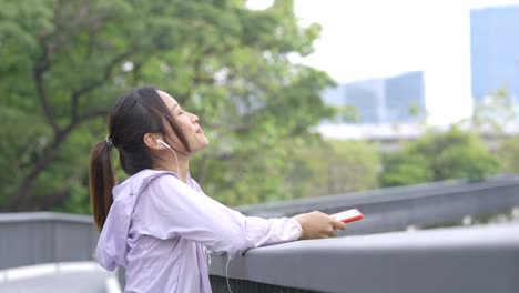 4k young asian woman listening to music from earphones and smartphone while jogging at public park the city in the morning.