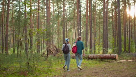 two sisters walking through lush green forest, one with blue hair tie and backpack, the other with cloth draped over bag, back view, with tall trees, soft light filtering through