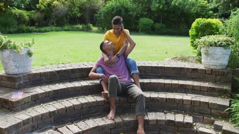 Smiling-mixed-race-gay-male-couple-sitting-on-steps-hugging-in-garden