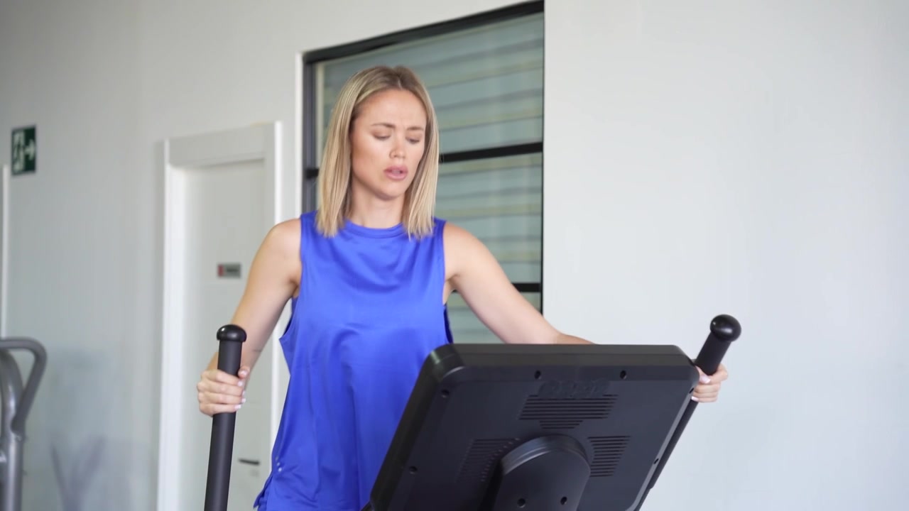 Una donna bionda si allena su una macchina ellittica in palestra, indossando un top a canotta blu.