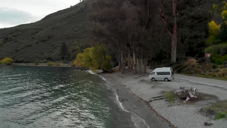 motorhome by beautiful blue lake wakatipu, queenstown, new zealand with mountains fresh snow cloudy sky in background - aerial drone