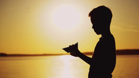 silhouette of a child by the river. paper boat is in the hands of the child. beautiful summer sunset. origami