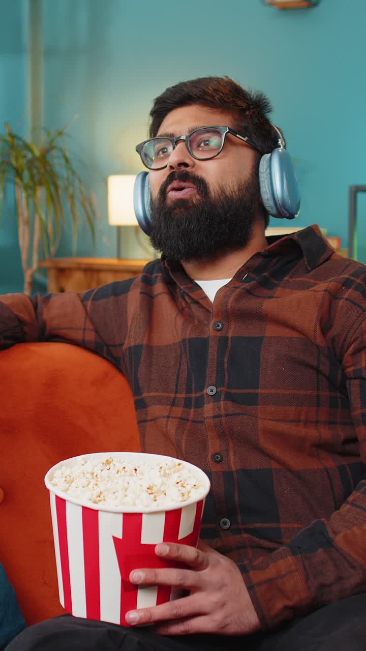 Homem árabe descontraído comendo pipoca, assistindo a um filme e aproveitando o fim de semana, sentado no sofá de casa