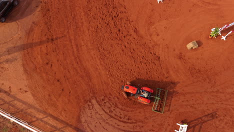Top-down aerial view of horseback riders training center with working tractor on red sandy arena.