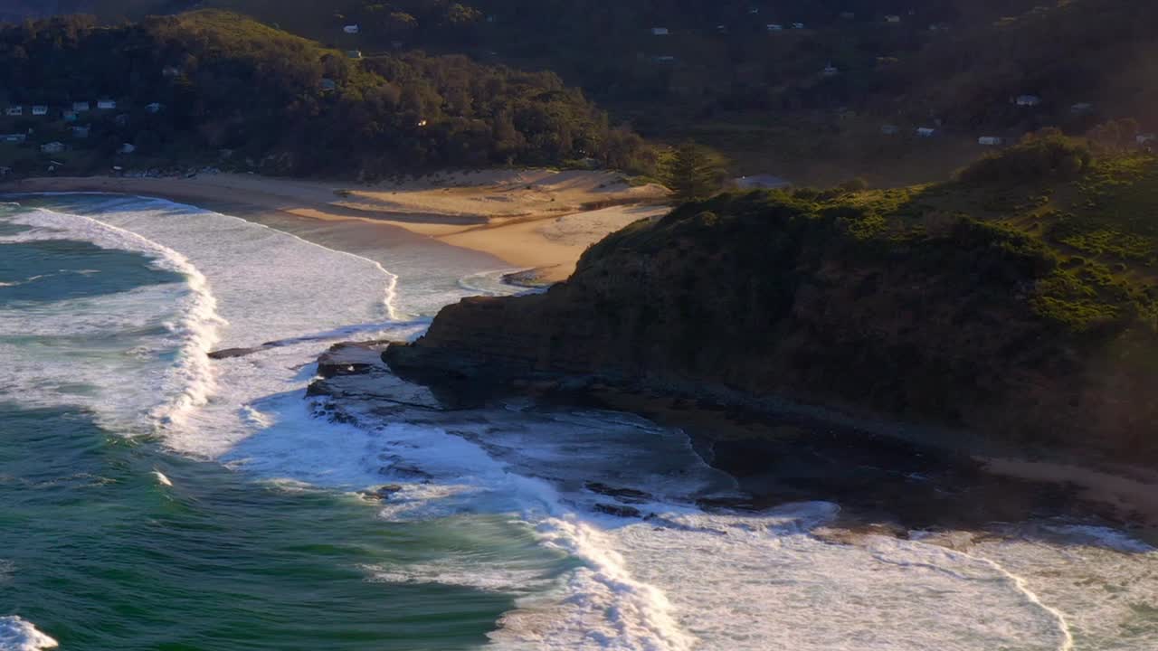 olas espumosas rodando hacia la costa de la era con cabañas en una exuberante meseta en el parque nacional real, nsw, australia