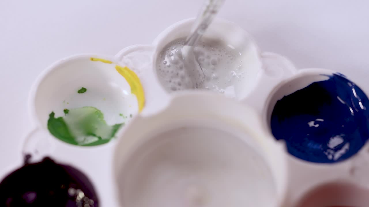 Paintbrush stirs white paint in plastic palette under bright lighting, top-down macro perspective