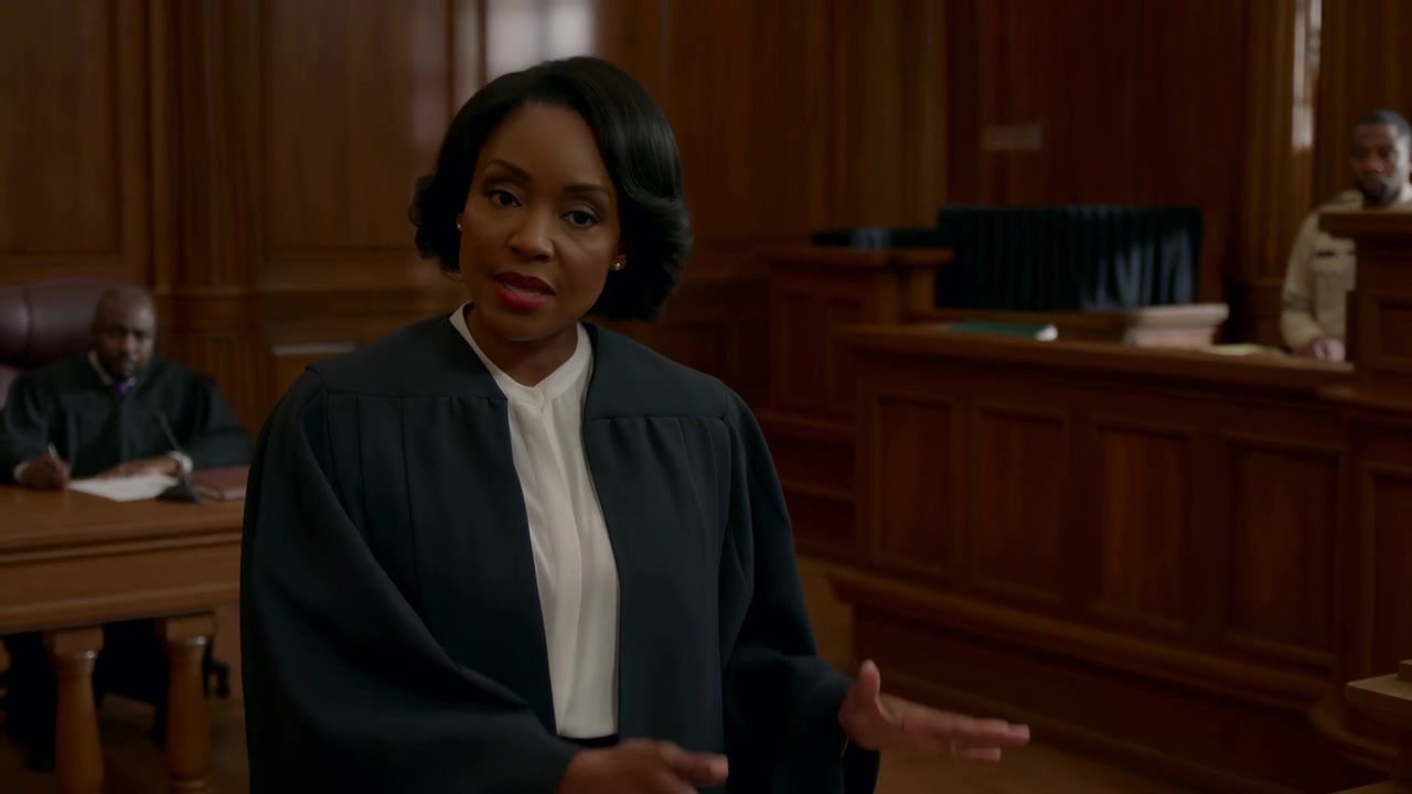 Uma mulher com uma toga escura está de pé em um tribunal com painéis de madeira, falando com a mão levantada enquanto homens sentados observam ao fundo sob luz interna quente.