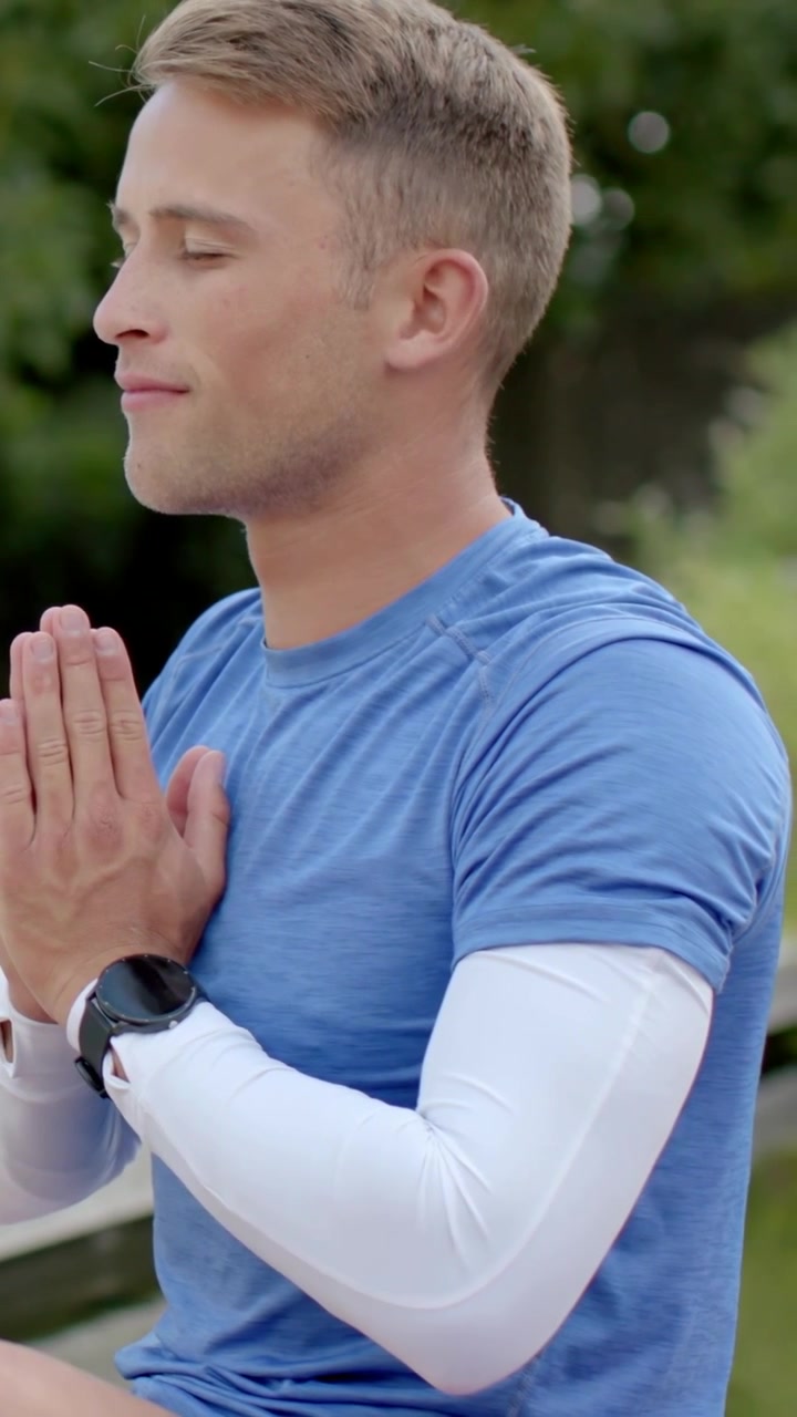Video verticale: Uomo che si rinfresca, indossa una maglietta blu, porta le mani al petto nel parco con uno smartwatch.
